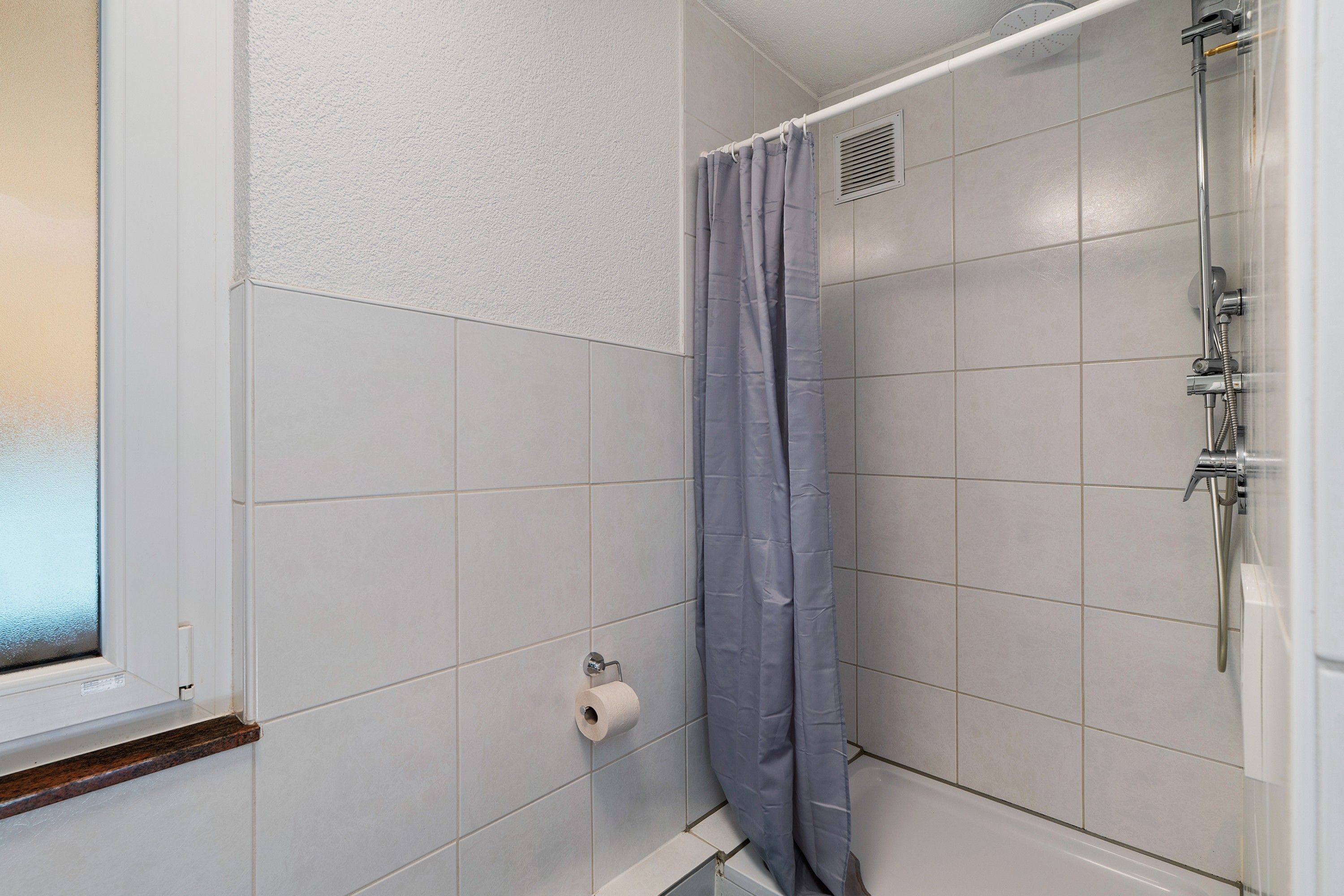 Ein Duschbereich mit grauer Duschvorhang, einer Handbrause und einer Toilettenpapierrolle an der Wand.