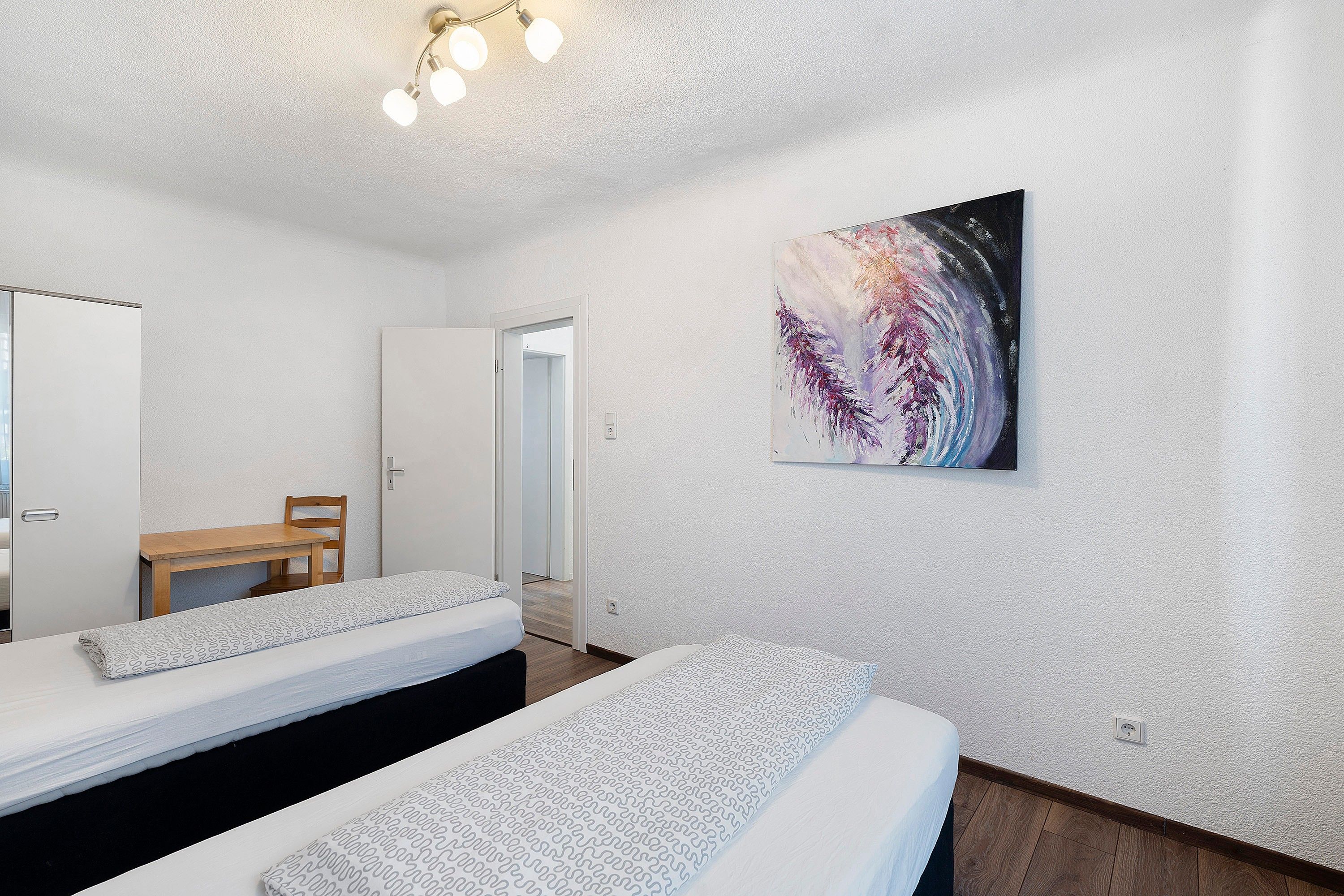 Een hotelkamer met twee bedden, een houten stoel, een schilderij aan de muur en een plafondlamp.