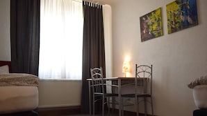 Quarto de hotel com uma cama, escrivaninha e duas cadeiras, radiador e duas pinturas na parede.