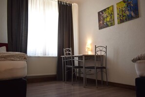 Habitación de hotel con una cama, un escritorio con dos sillas, un radiador y dos cuadros en la pared.