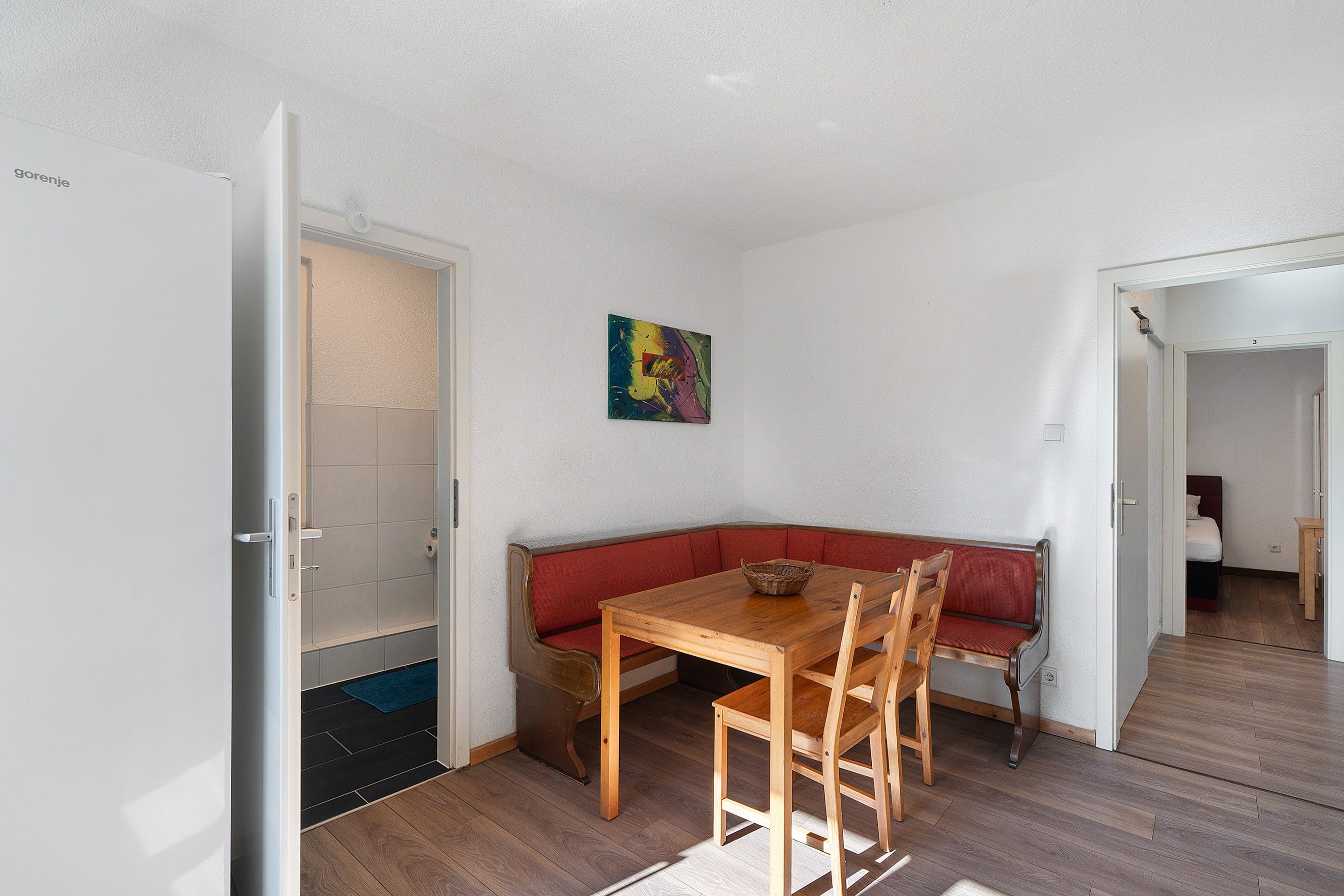 Een kamer met een houten tafel, een bankje, een deur en een kleurrijk abstract schilderij aan de muur.