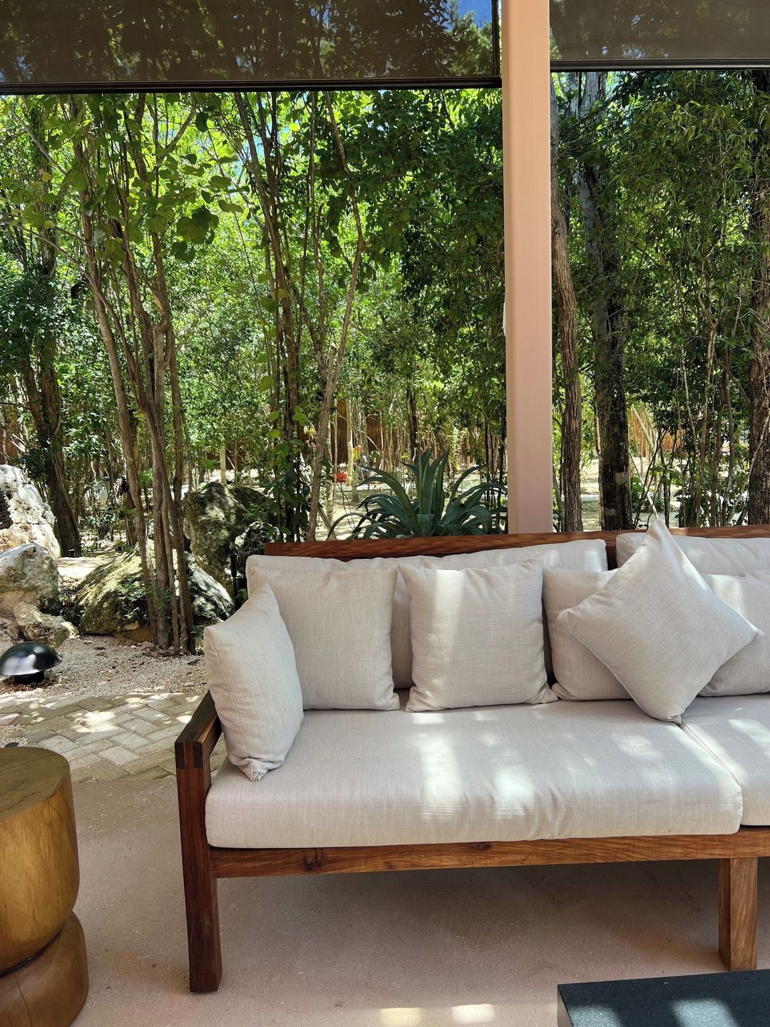 A wooden sofa with cushions in a room with large windows overlooking a forested area.