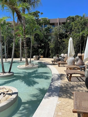 Área de piscina com espreguiçadeiras e guarda-sóis, cercada por palmeiras e um edifício ao fundo.