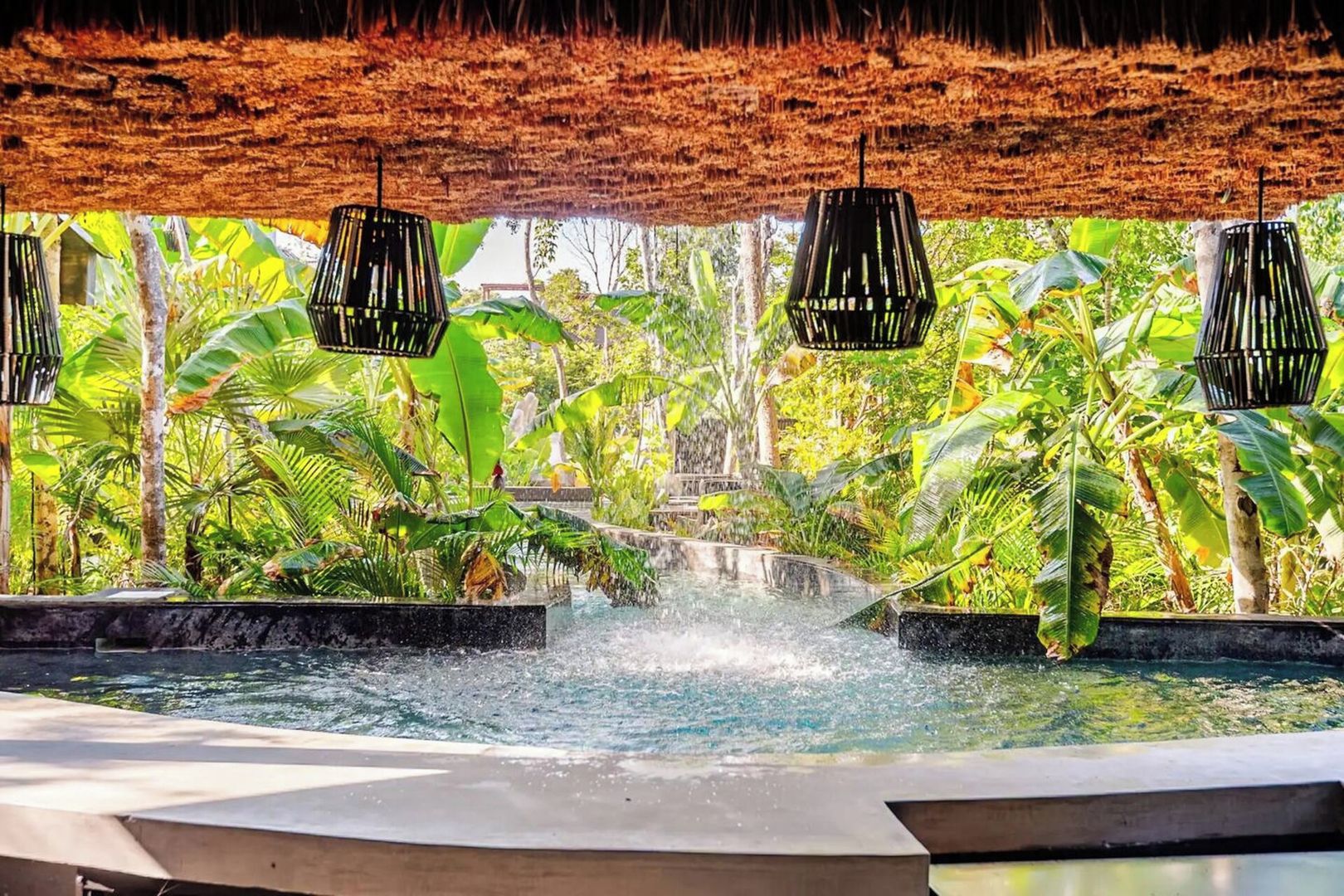 A pool with a waterfall feature surrounded by lush greenery and thatched roof structures.