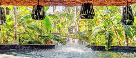 A pool with a waterfall feature surrounded by lush greenery and thatched roof structures.