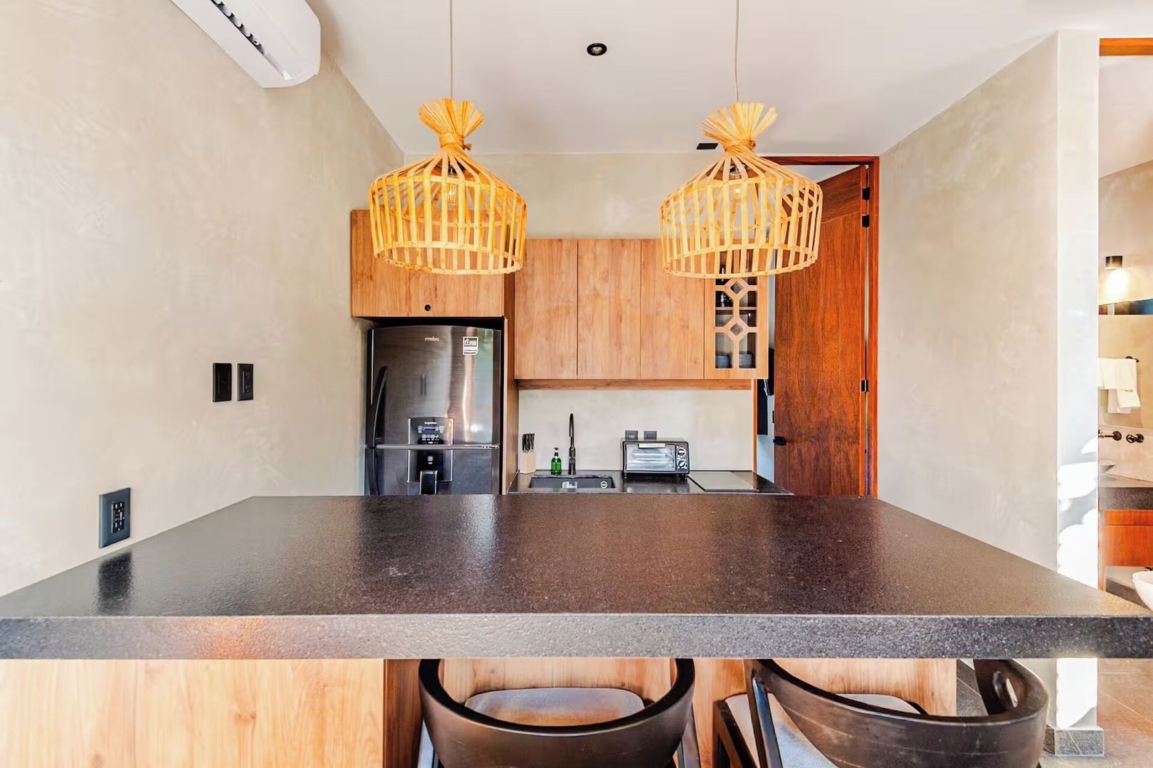 A modern kitchen with a central island, wooden cabinets, a stainless steel refrigerator, and two hanging light fixtures.