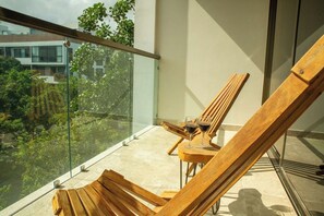 A balcony with wooden lounge chairs and a table, a glass railing, and a view of greenery and buildings. - Please note: electricity is not included in reservations longer than 28... (Tulum)