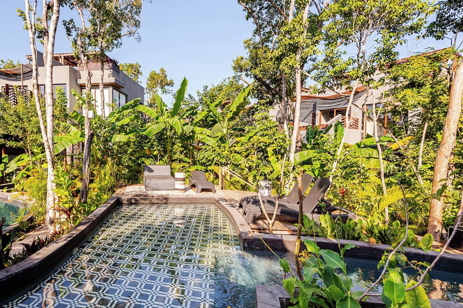 Un espace piscine doté de chaises longues et d’une terrasse carrelée, le tout entouré d’une végétation tropicale.