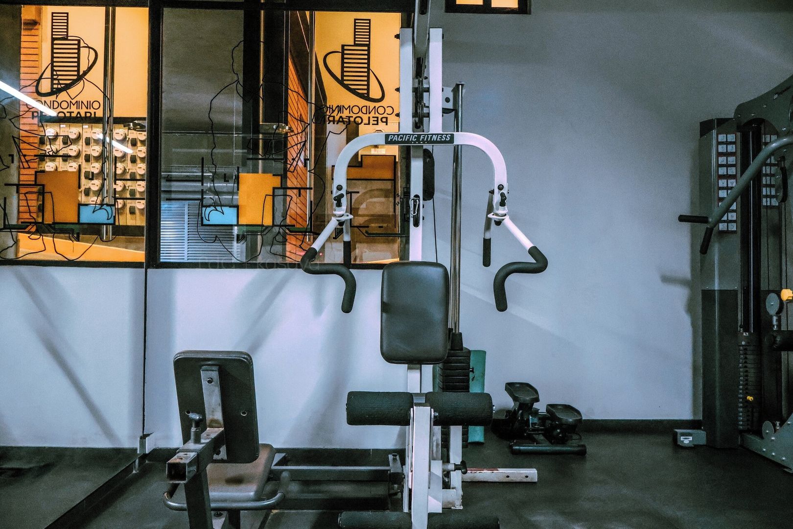 A gym with exercise equipment, a window with a view of buildings, and a sign that reads 'PACIFIC FITNESS'.