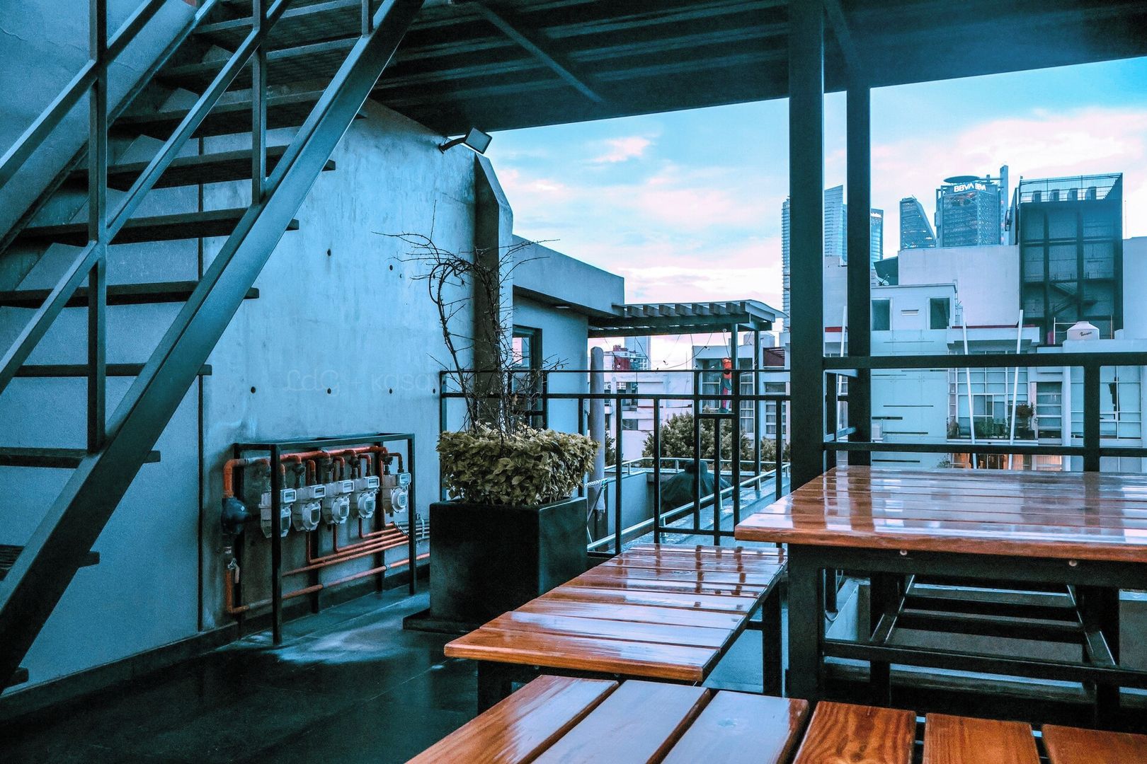 A rooftop terrace with a staircase, a seating area with wooden tables, and a view of city buildings.