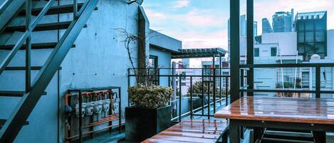 A rooftop terrace with a staircase, a seating area with wooden tables, and a view of city buildings.