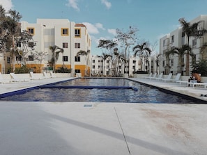 Ein Swimmingpool mit Liegestühlen und Palmen, umgeben von mehrstöckigen Gebäuden.