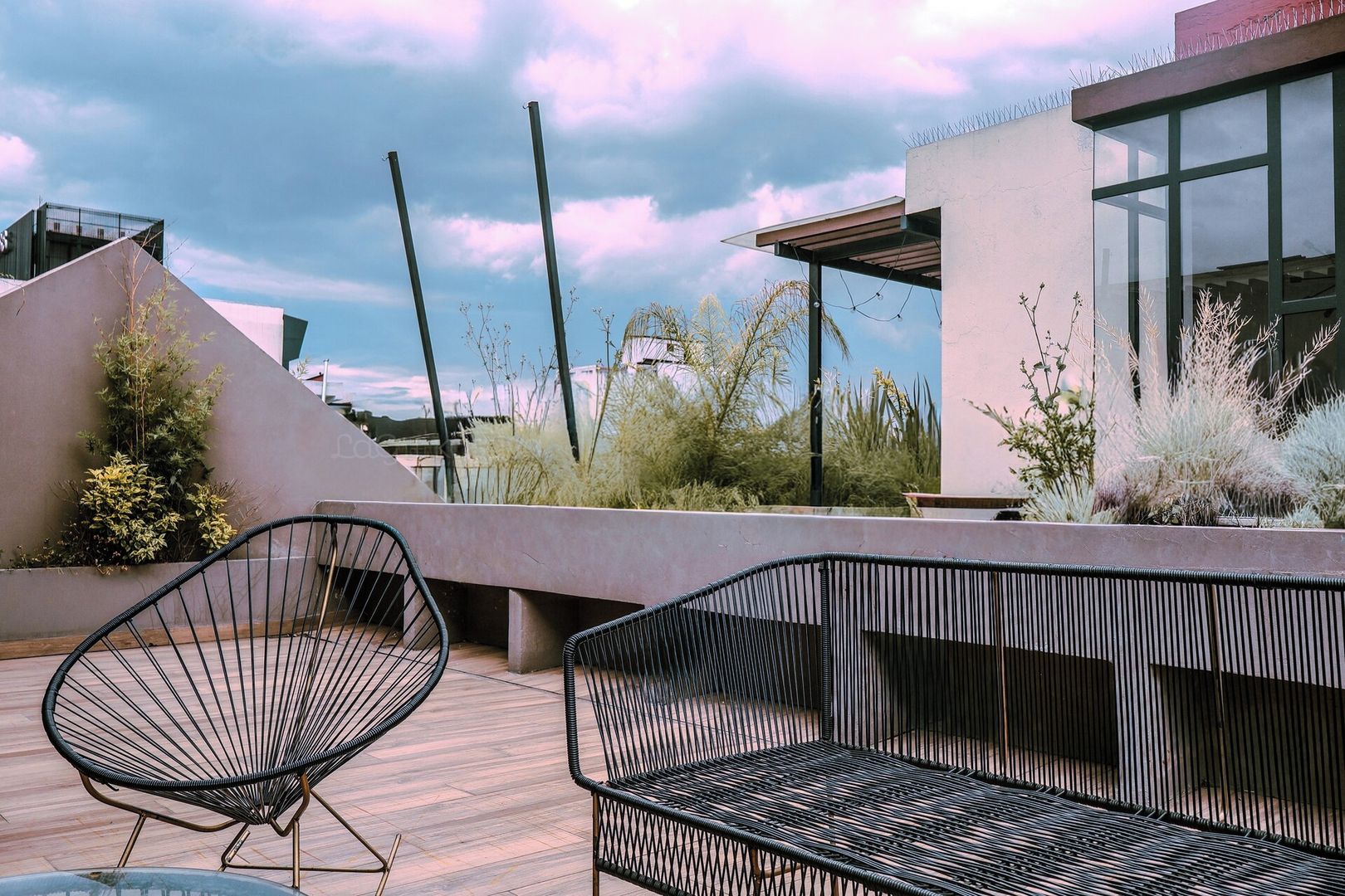 A rooftop terrace with two black wireframe chairs, a concrete bench, and a glass table.