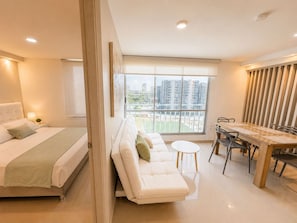 A modern hotel room with a bed, sofa, dining table, chairs, and a large window with a city view. - Palmar Apartment – Near Buenavista by SOHO – 34A (Barranquilla)