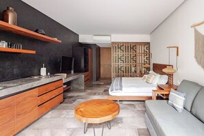 A modern hotel room with a kitchen area, a bed with a wooden headboard, a grey sofa, and a round wooden table. - Premium studio · Private immersion · Gym and Sauna (Tulum)