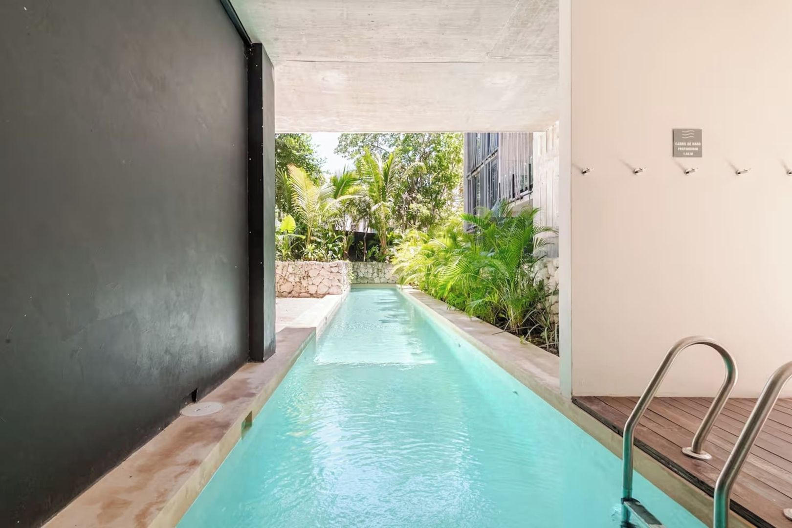 A swimming pool with a stone wall and metal railings, surrounded by greenery and a building in the background.