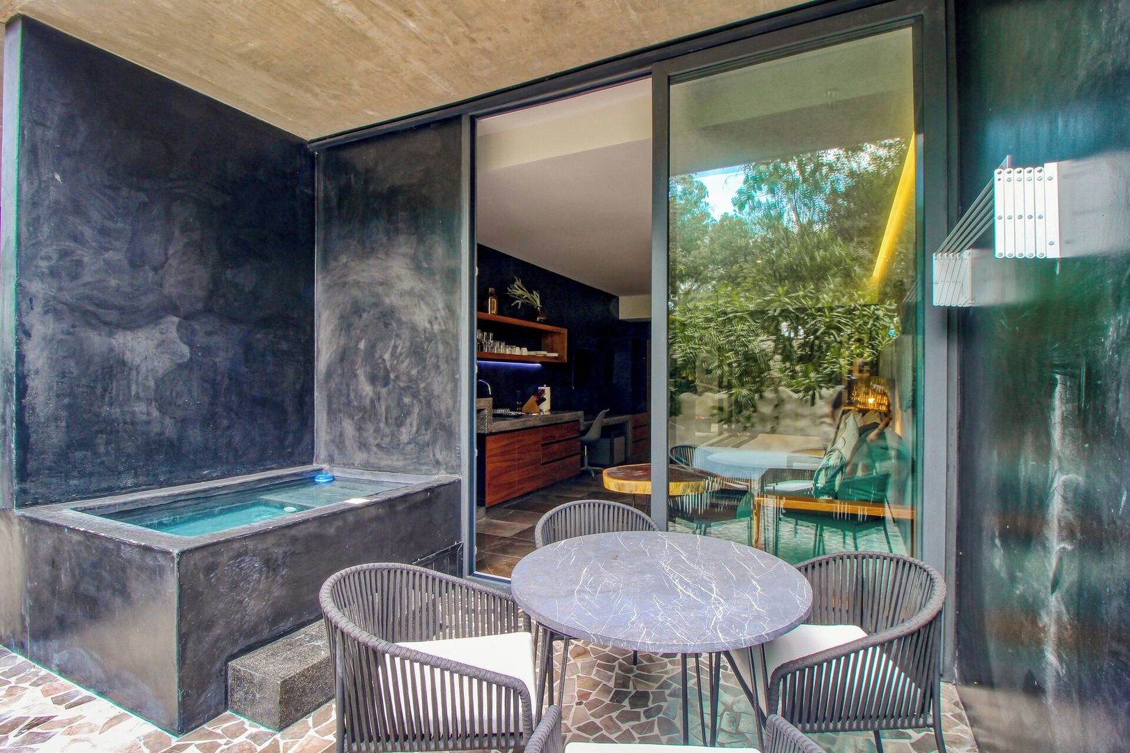 A modern outdoor seating area with a pool, wicker chairs, and a glass-enclosed space with a view of greenery.