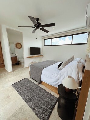 A bedroom with a bed, a ceiling fan, a TV on a shelf, a window, and a door leading to a bathroom. - PH con alberca privada (Tulum)