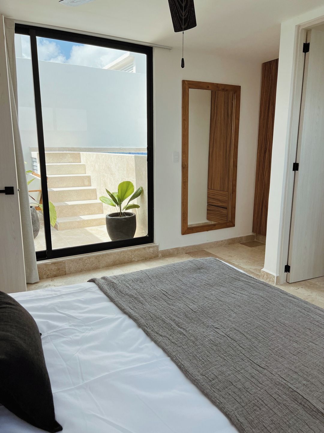 A bedroom with a bed, a potted plant, a sliding glass door leading to a balcony, and a wooden wardrobe.