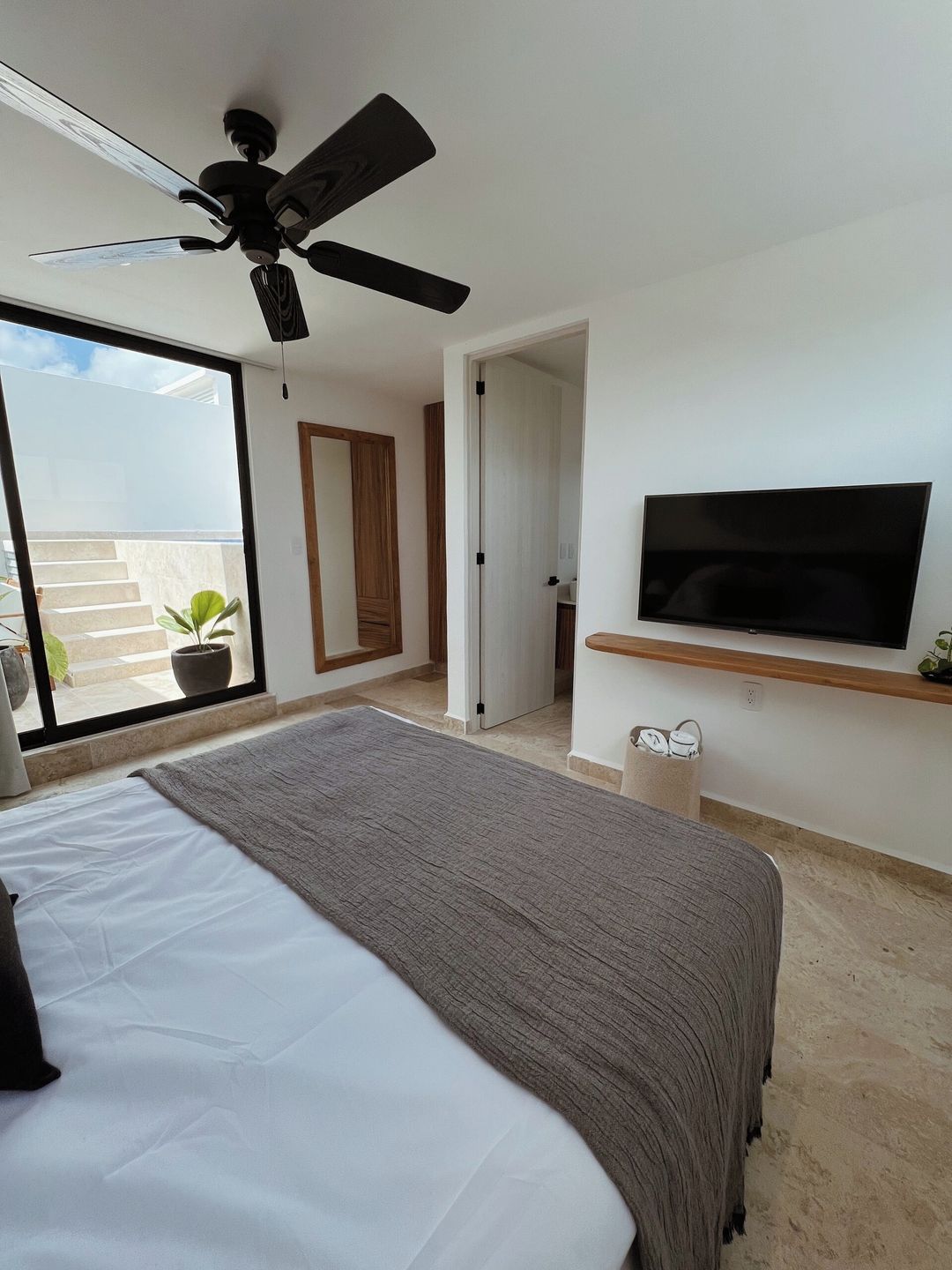 A bedroom with a bed, a ceiling fan, a TV mounted on the wall, and a balcony with potted plants.