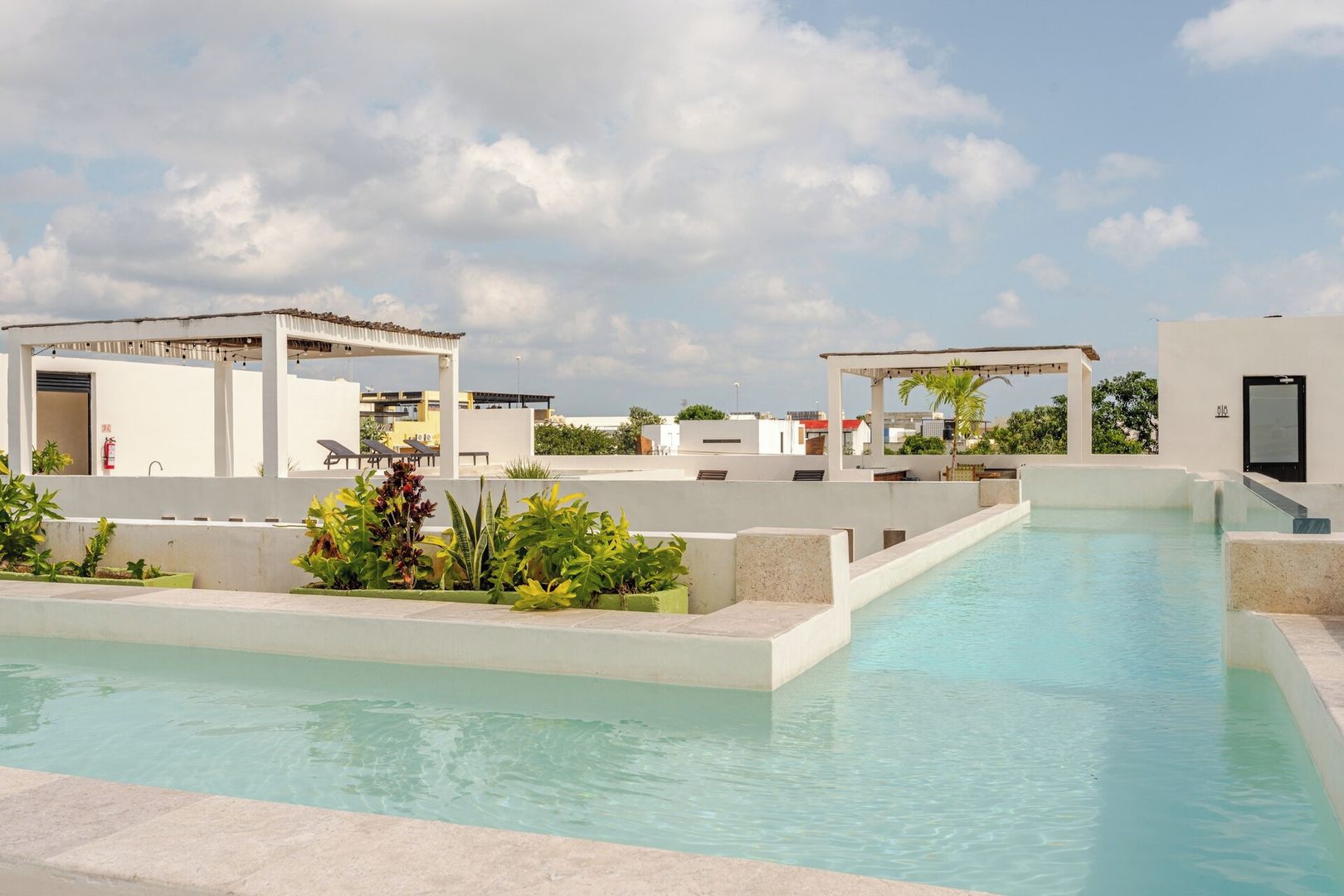 Una piscina en la azotea con cielo despejado, rodeada de estructuras blancas y follaje.