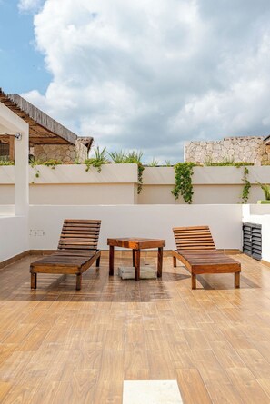 A rooftop terrace with wooden lounge chairs, a small table, and a stone wall with greenery. - Stylish Tulum Apt w/Jacuzzi and Pools (Tulum)