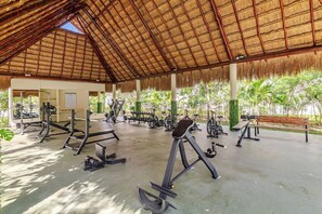 Een buitengymzaal met trainingsapparatuur, een rieten dak en uitzicht op palmbomen.