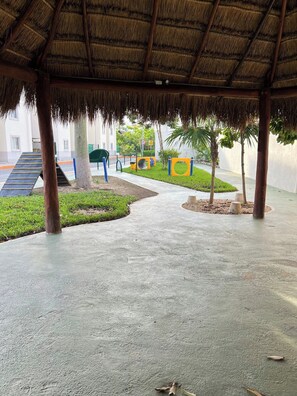 A covered outdoor area with a concrete floor, a thatched roof, and a playground with slides and climbing structures.