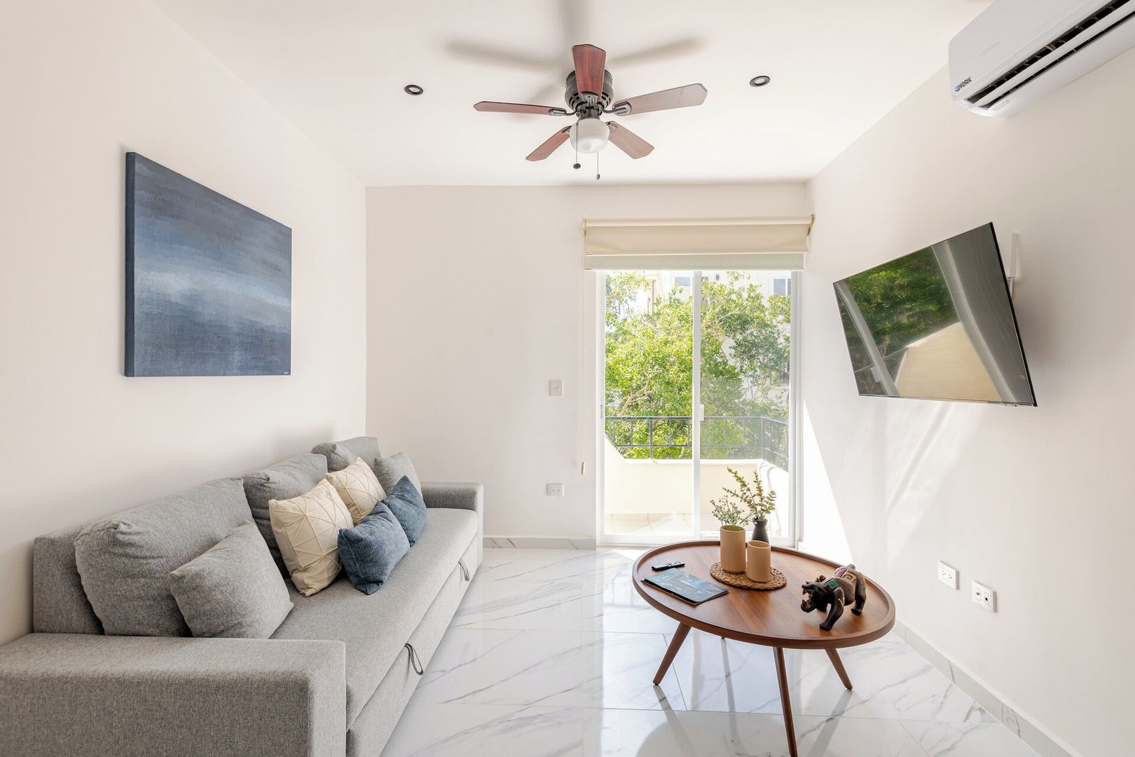 Un salon moderne avec un canapé gris, un ventilateur de plafond, un téléviseur à écran plat et une grande fenêtre donnant sur un paysage verdoyant.