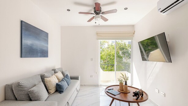 Ein modernes Wohnzimmer mit einem grauen Sofa, einem Deckenventilator, einem Flachbildfernseher und einem großen Fenster mit Blick auf Grünflächen.