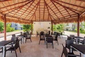 Un patio al aire libre con techo de paja, amueblado con mesas y sillas, y una zona de barbacoa.