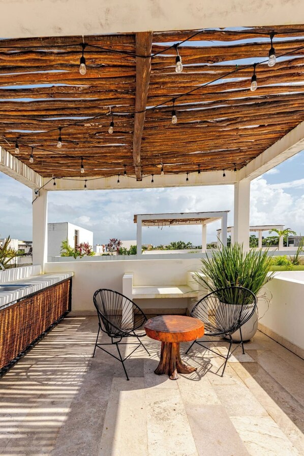 An outdoor patio with a wooden pergola, two black wireframe chairs, a round table, and a potted plant. - Modern Apartment with Jacuzzi (Tulum)
