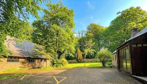 A paved driveway leading to a building with a brick facade and large windows, surrounded by lush greenery and trees. - Charming 1-bedroom cottage on 33 acre woodland estate with hot tub & log burner (Hollybush)