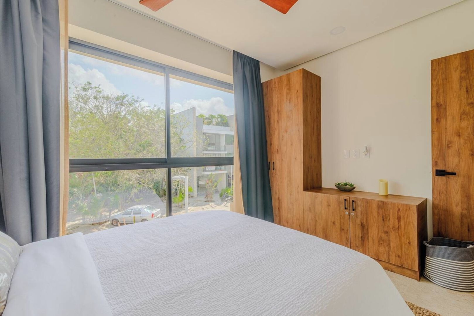 Une chambre avec une grande fenêtre, un lit, une armoire en bois et une petite plante posée sur la commode.
