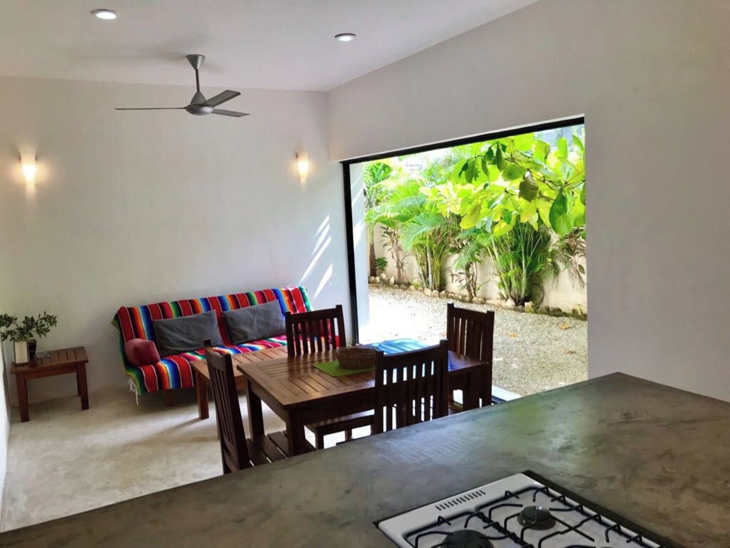 Un intérieur moderne avec un canapé, une table à manger, des chaises, un ventilateur de plafond et une vue sur la verdure à l’extérieur.