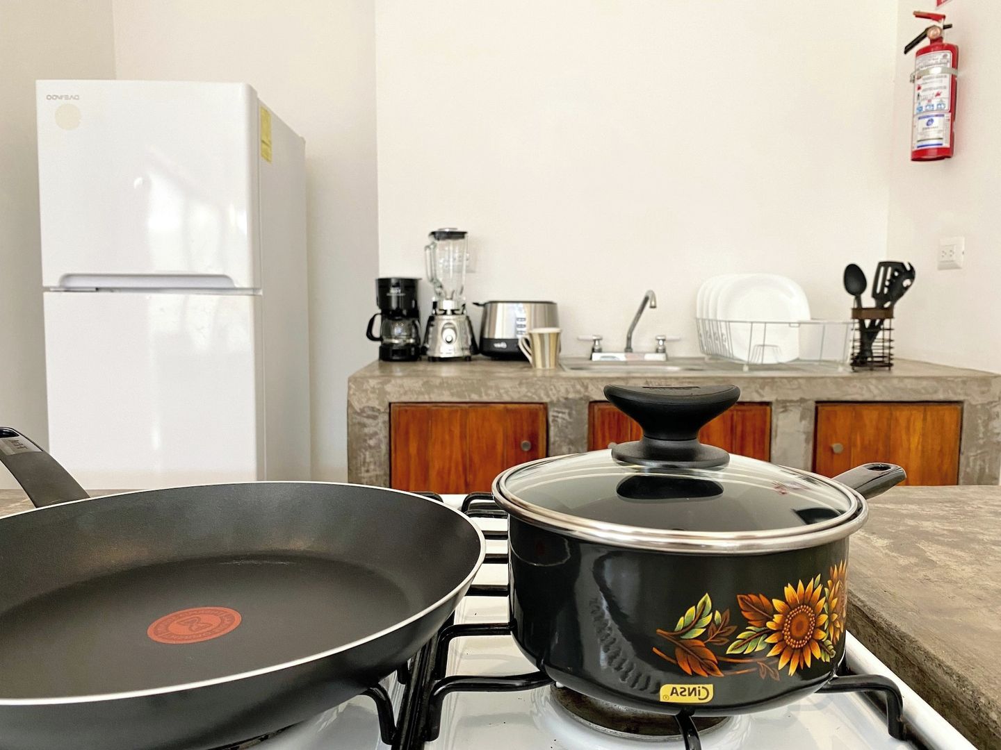A kitchen with a stove, a frying pan, a pot with a floral design, a coffee maker, and a white refrigerator.