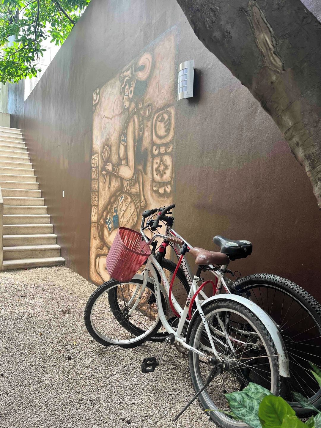 A bicycle parked against a wall with a mural, a red basket attached to the bike, and a staircase leading up.