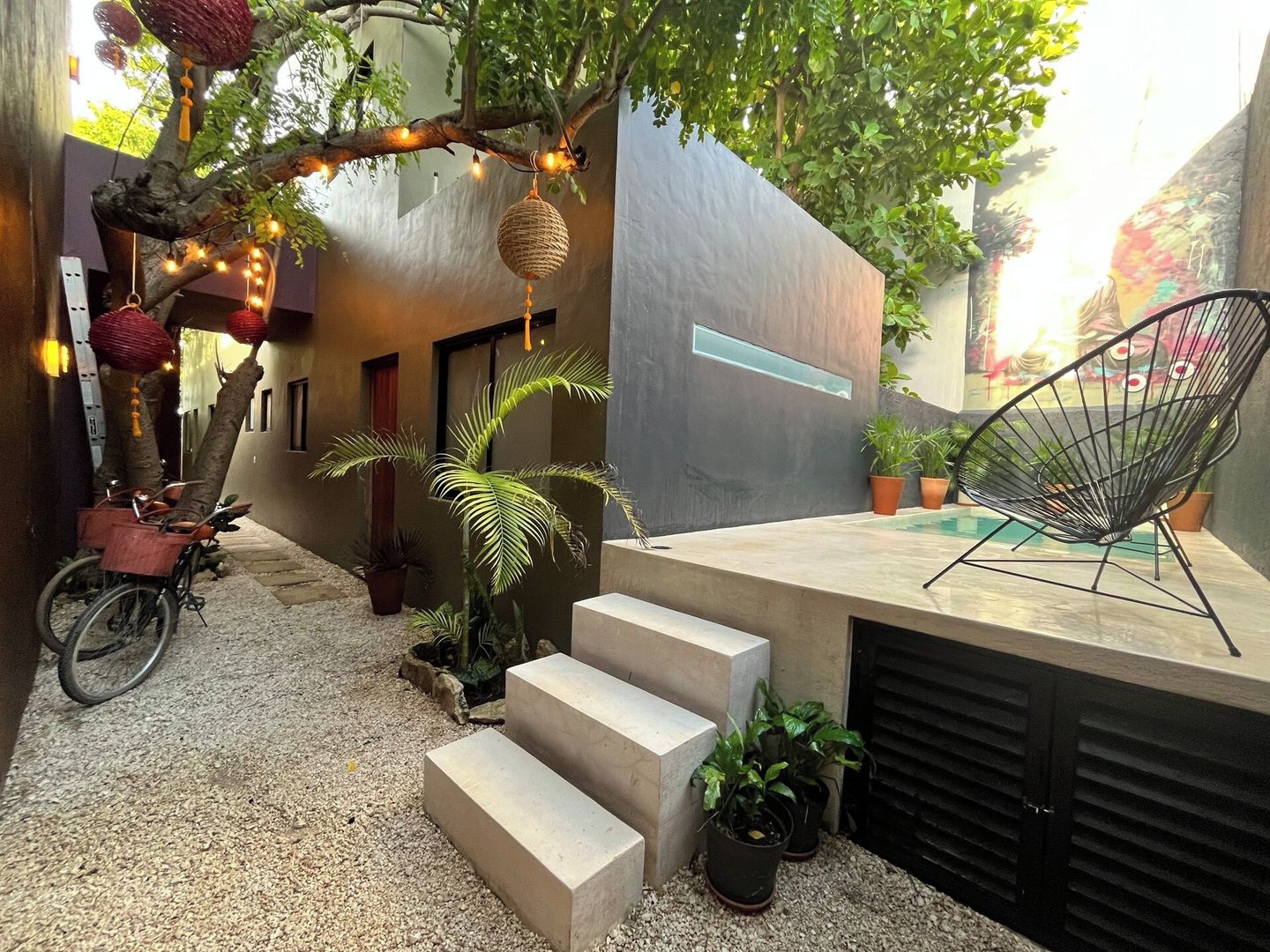 A modern outdoor space with a concrete bench, a bicycle, potted plants, and a tree with hanging lights.