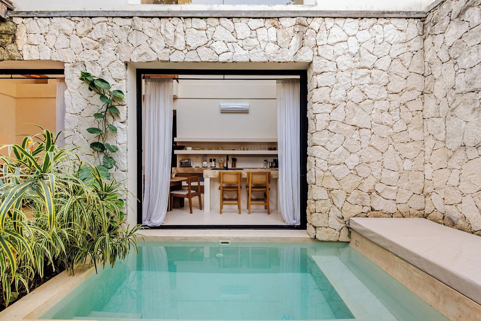 A pool area with a stone wall, a kitchen visible through an open door, and potted plants.