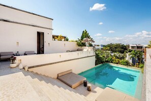 Ein moderner Poolbereich im Freien mit einem klaren, blauen Pool, einer Lounge-Ecke mit Kissen und einer Treppe zu einer überdachten Terrasse.
