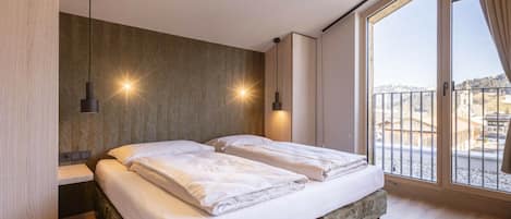 A bedroom with a bed, two bedside lamps, a window with a view, and a wooden floor.