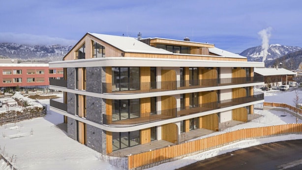 Un edificio moderno de varios pisos con amplios ventanales y balcones, situado frente a un paisaje nevado con montañas al fondo.