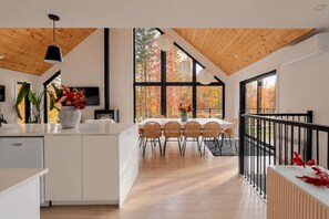A modern kitchen with a central island, dining area with chairs, and a view of autumn trees through large windows.