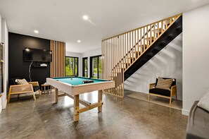 A modern living room with a pool table, a staircase, and a TV mounted on the wall.