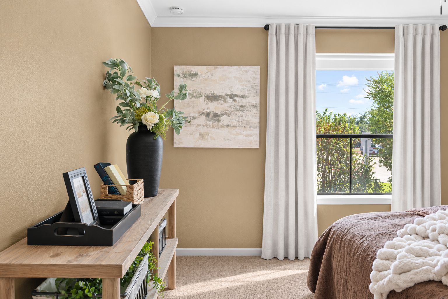 A bedroom with a large window, a bed with a white blanket, a wooden console table with books and a vase, and a painting on the wall.