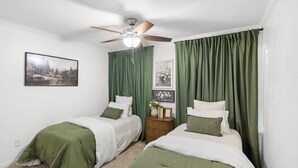 Een slaapkamer met twee bedden, groene gordijnen, een plafondventilator en een schilderij aan de muur.