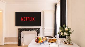 Une pièce avec un téléviseur affichant le logo « NETFLIX », une cheminée, une table à manger dressée avec de la nourriture et des boissons, et un vase rempli de fleurs.