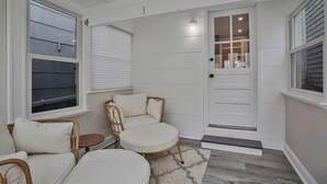 Una habitación con una puerta blanca, una silla de mimbre, una mesa redonda y una alfombra pequeña.
