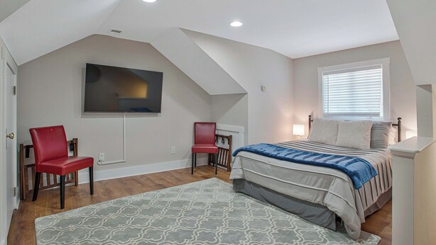 Un dormitorio con techo inclinado, una cama con colcha a rayas, dos sillas rojas, un televisor montado en la pared y una ventana con persianas.