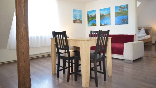 A dining area with a wooden table and chairs, a red sofa, and a wall with landscape paintings. - Holiday Home Neuhausen – Attic Apartment / 2 Bedrooms / Wi-Fi (Neuhausen)